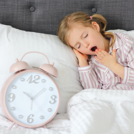 Child yawning in bed beside a clock